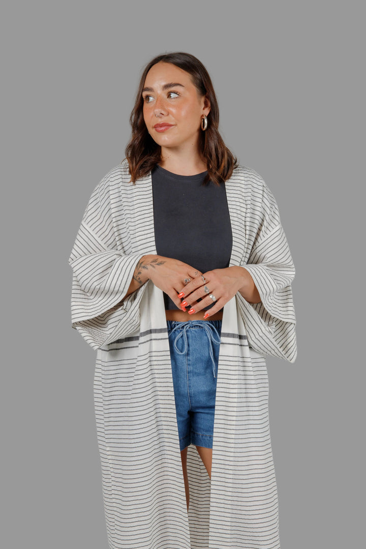 Woman wearing a long, lightweight off white striped open cover-up/robe with grey stripes and side slits over casual clothing on a plain background