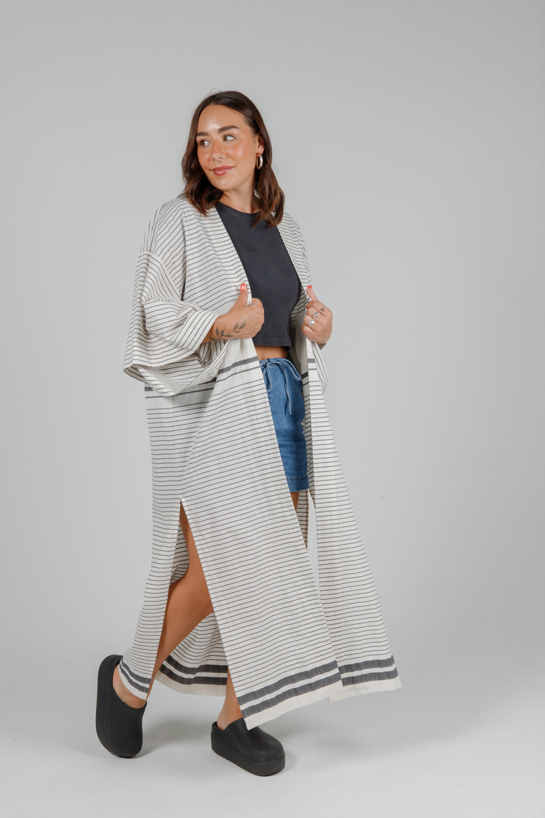 Woman wearing a long, lightweight off white striped open cover-up/robe with grey stripes and side slits over casual clothing on a plain background