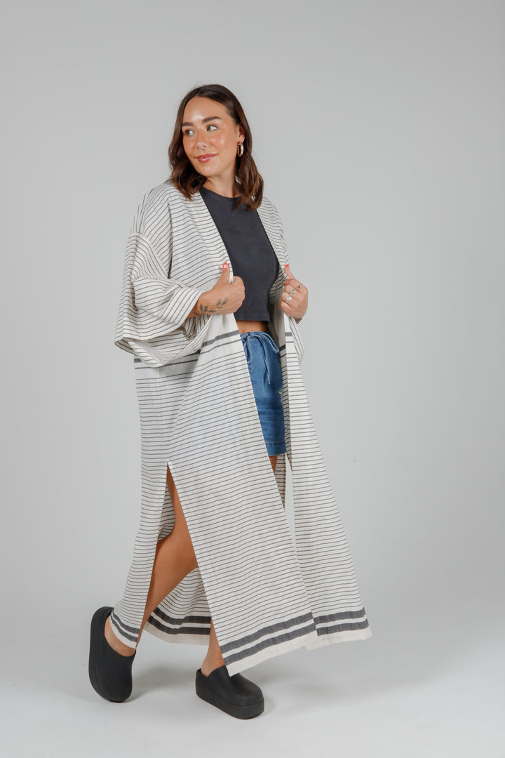 Woman wearing a long, lightweight off white striped open cover-up/robe with grey stripes and side slits over casual clothing on a plain background