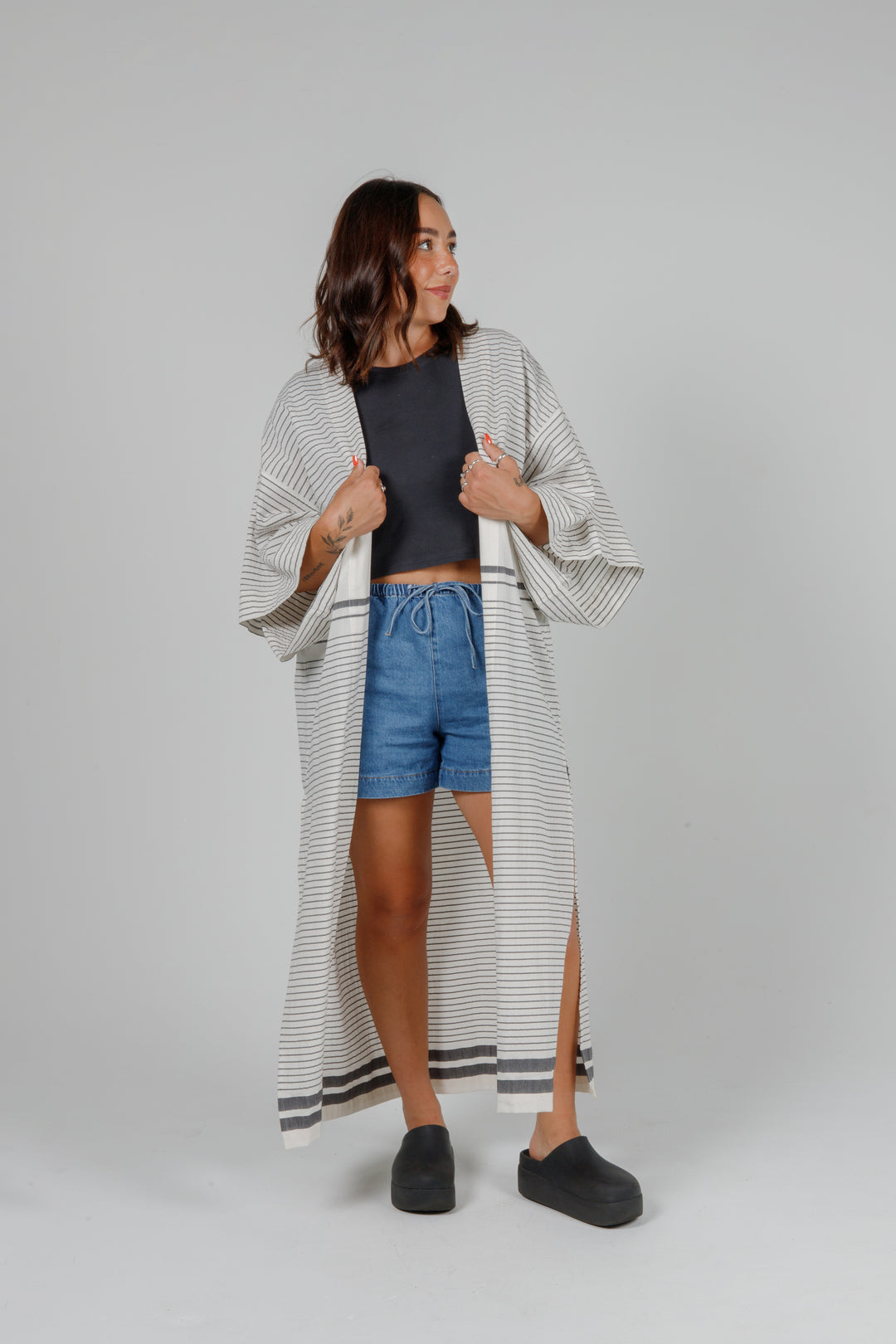 Woman wearing a long, lightweight off white striped open cover-up/robe with grey stripes and side slits over casual clothing on a plain background