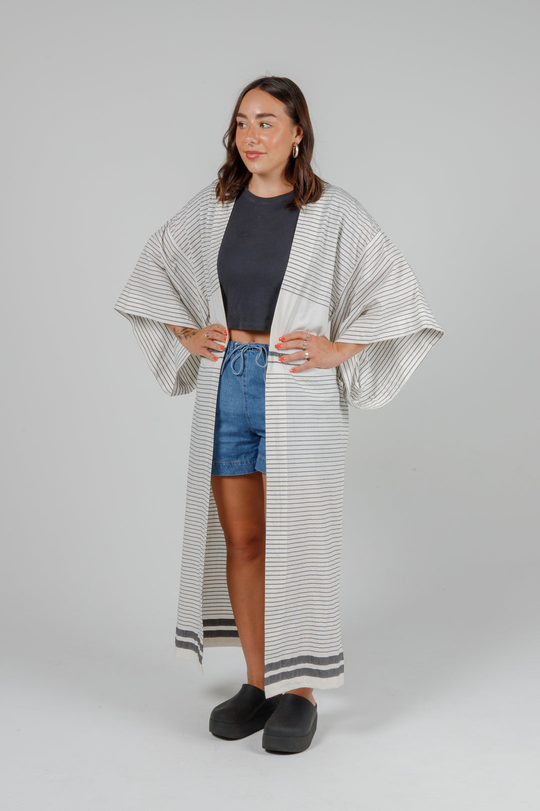 Woman wearing a long, lightweight off white striped open cover-up/robe with grey stripes and side slits over casual clothing on a plain background
