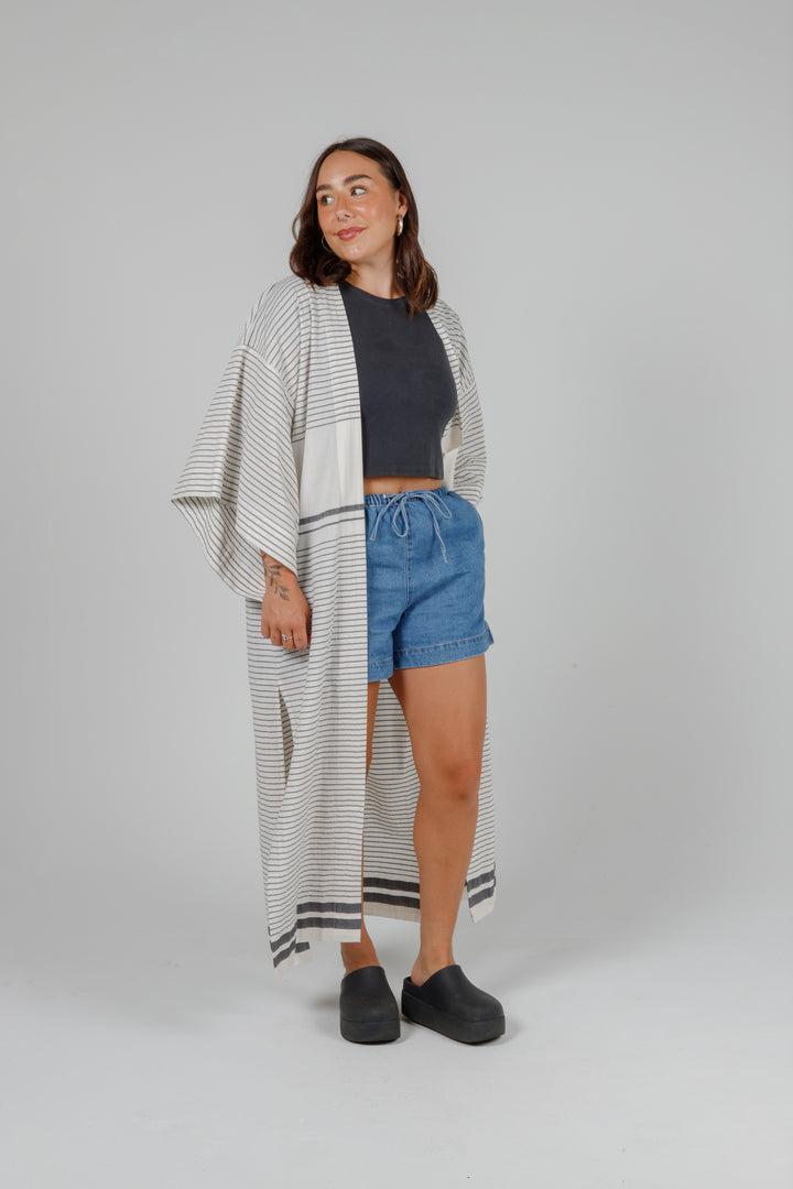 Woman wearing a long, lightweight off white striped open cover-up/robe with grey stripes and side slits over casual clothing on a plain background