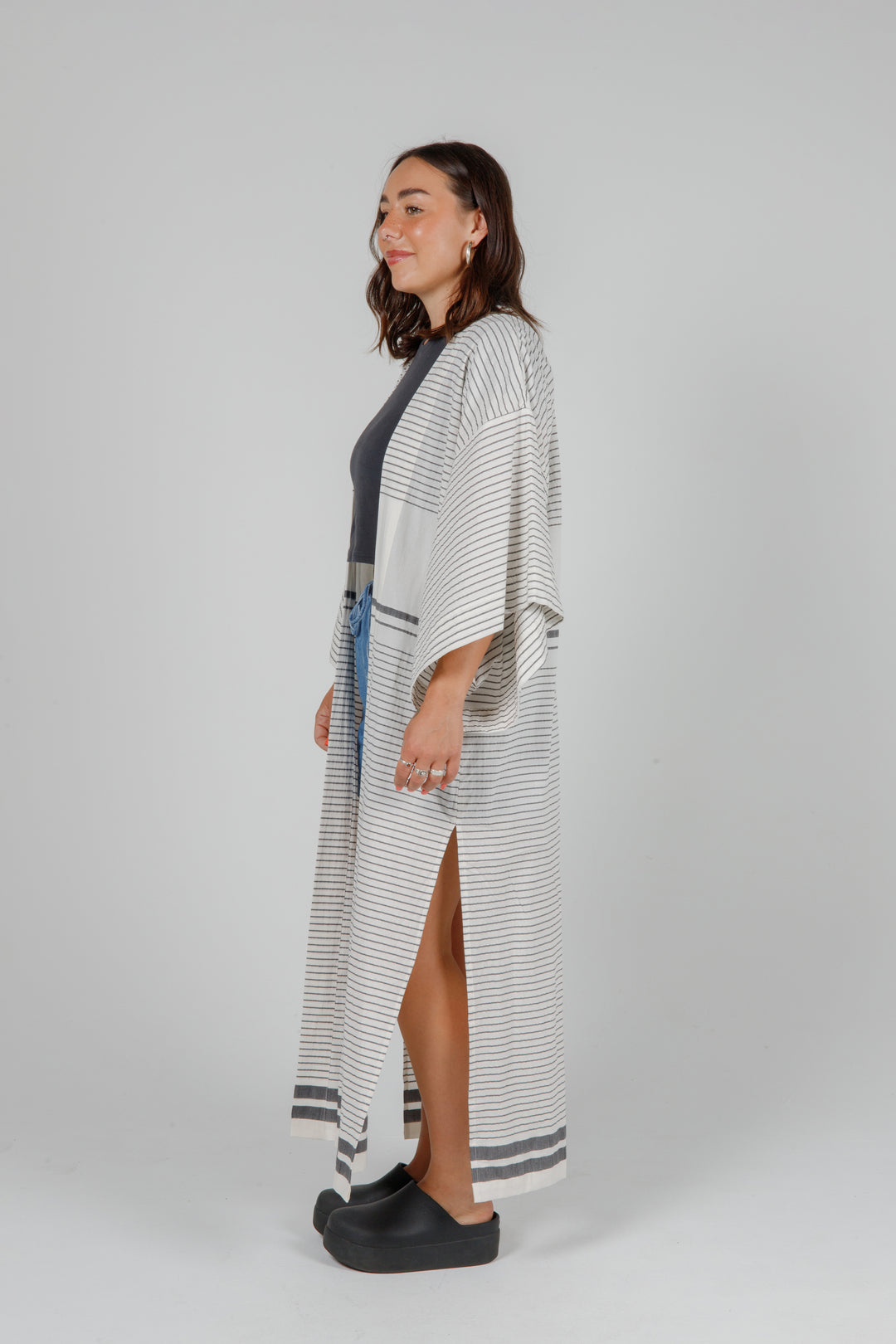 Woman wearing a long, lightweight off white striped open cover-up/robe with grey stripes and side slits over casual clothing on a plain background
