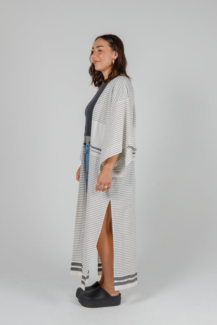 Woman wearing a long, lightweight off white striped open cover-up/robe with grey stripes and side slits over casual clothing on a plain background