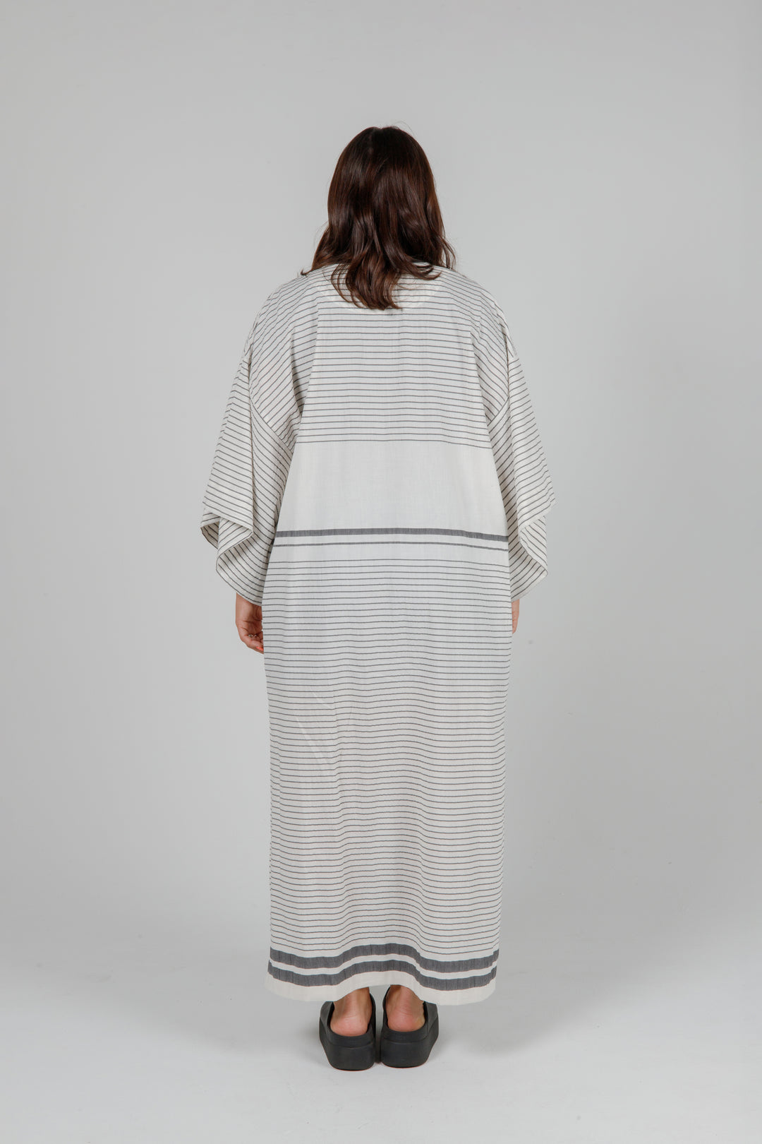 Woman wearing a long, lightweight off white striped open cover-up/robe with grey stripes and side slits over casual clothing on a plain background