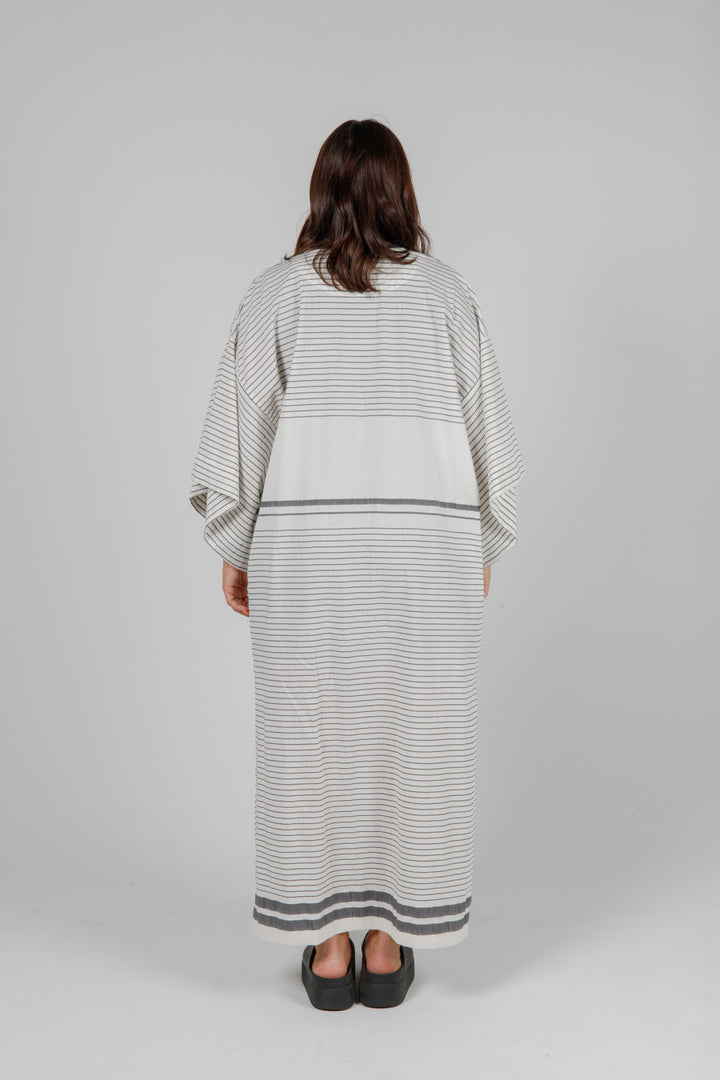 Woman wearing a long, lightweight off white striped open cover-up/robe with grey stripes and side slits over casual clothing on a plain background