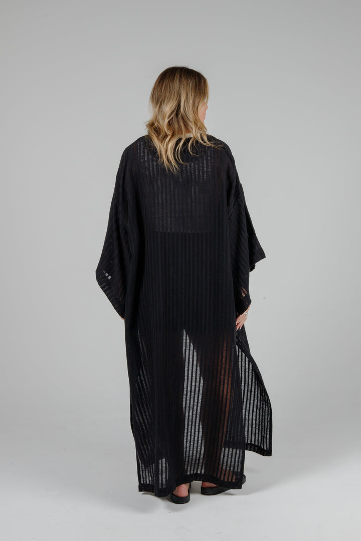 View of back of Long black semi-sheer linen blend cover-up with wide kimono sleeves and side slits, full-length lightweight beach kimono layered over neutral outfit for resort wear style