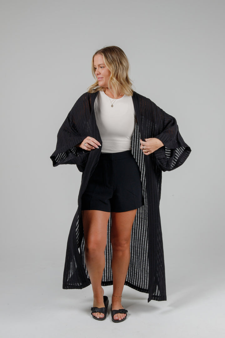 Woman wearing a black long-sleeve top and black shorts front view of Long black semi-sheer linen blend cover-up with wide kimono sleeves and side slits, full-length lightweight beach kimono layered over neutral outfit for resort wear style