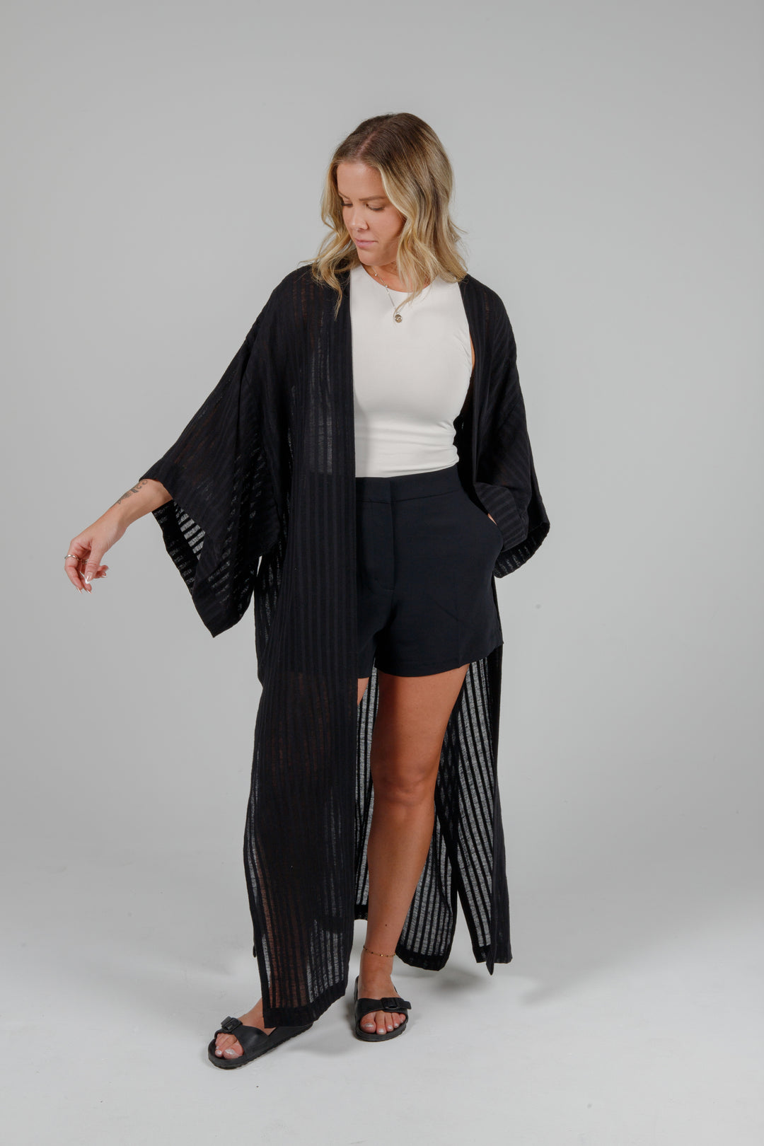 Woman wearing a black kimono-style garment over a white top and black shorts Long black semi-sheer linen blend cover-up with wide kimono sleeves and side slits, full-length lightweight beach kimono layered over neutral outfit for resort wear style