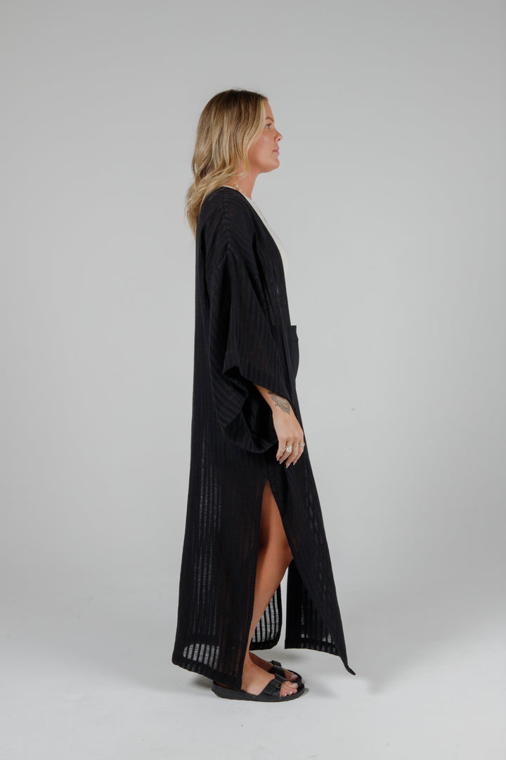 Side view of Long black semi-sheer linen blend cover-up with wide kimono sleeves and side slits, full-length lightweight beach kimono layered over neutral outfit for resort wear style