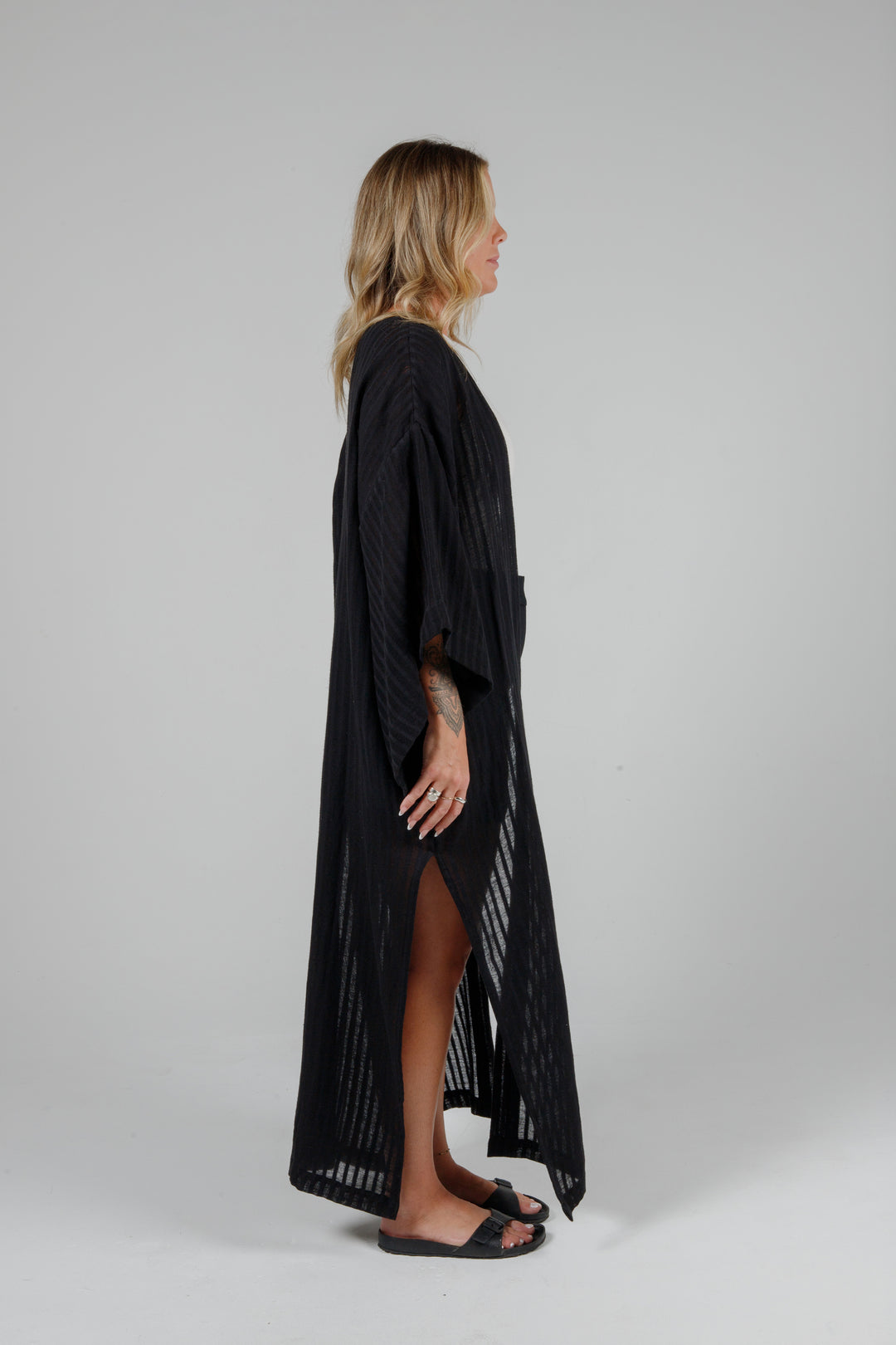 side view of Long black semi-sheer linen blend cover-up with wide kimono sleeves and side slits, full-length lightweight beach kimono layered over neutral outfit for resort wear style
