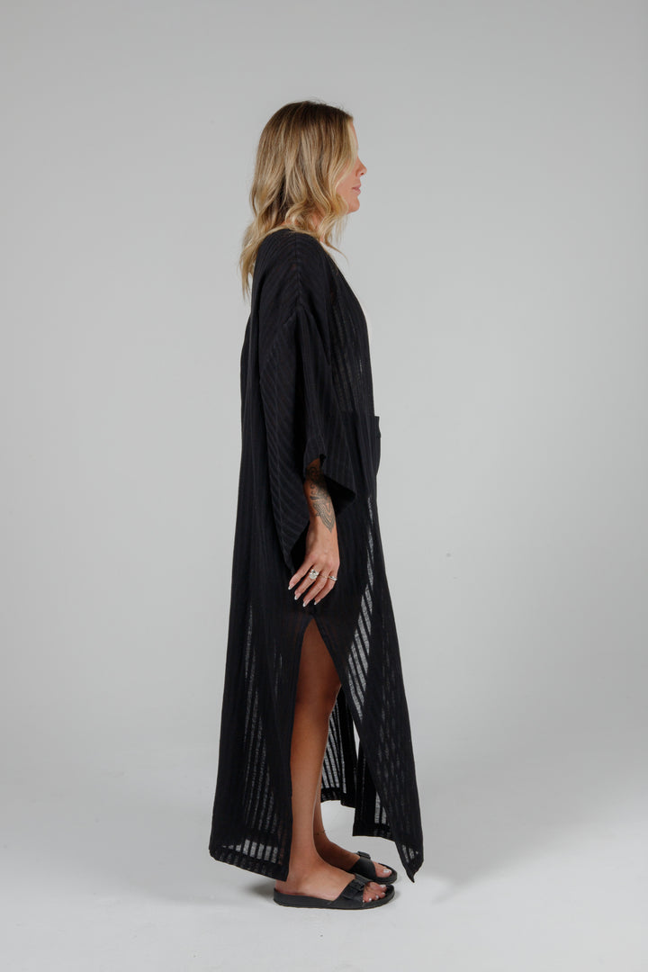 side view of Long black semi-sheer linen blend cover-up with wide kimono sleeves and side slits, full-length lightweight beach kimono layered over neutral outfit for resort wear style