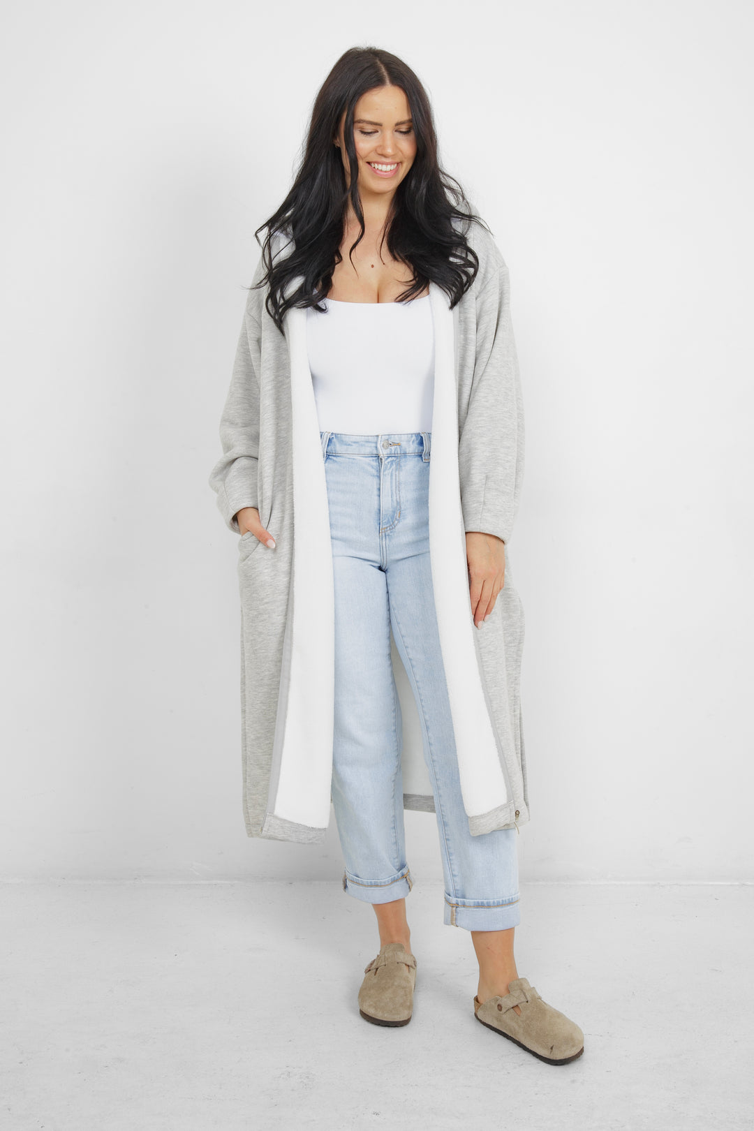 Woman wearing heather grey a long zip up hoodie, unzipped with side slits and pockets in front of a plain grey background