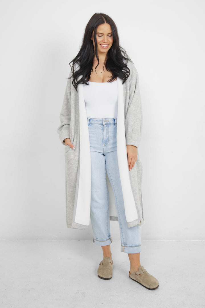 Woman wearing heather grey a long zip up hoodie, unzipped with side slits and pockets in front of a plain grey background