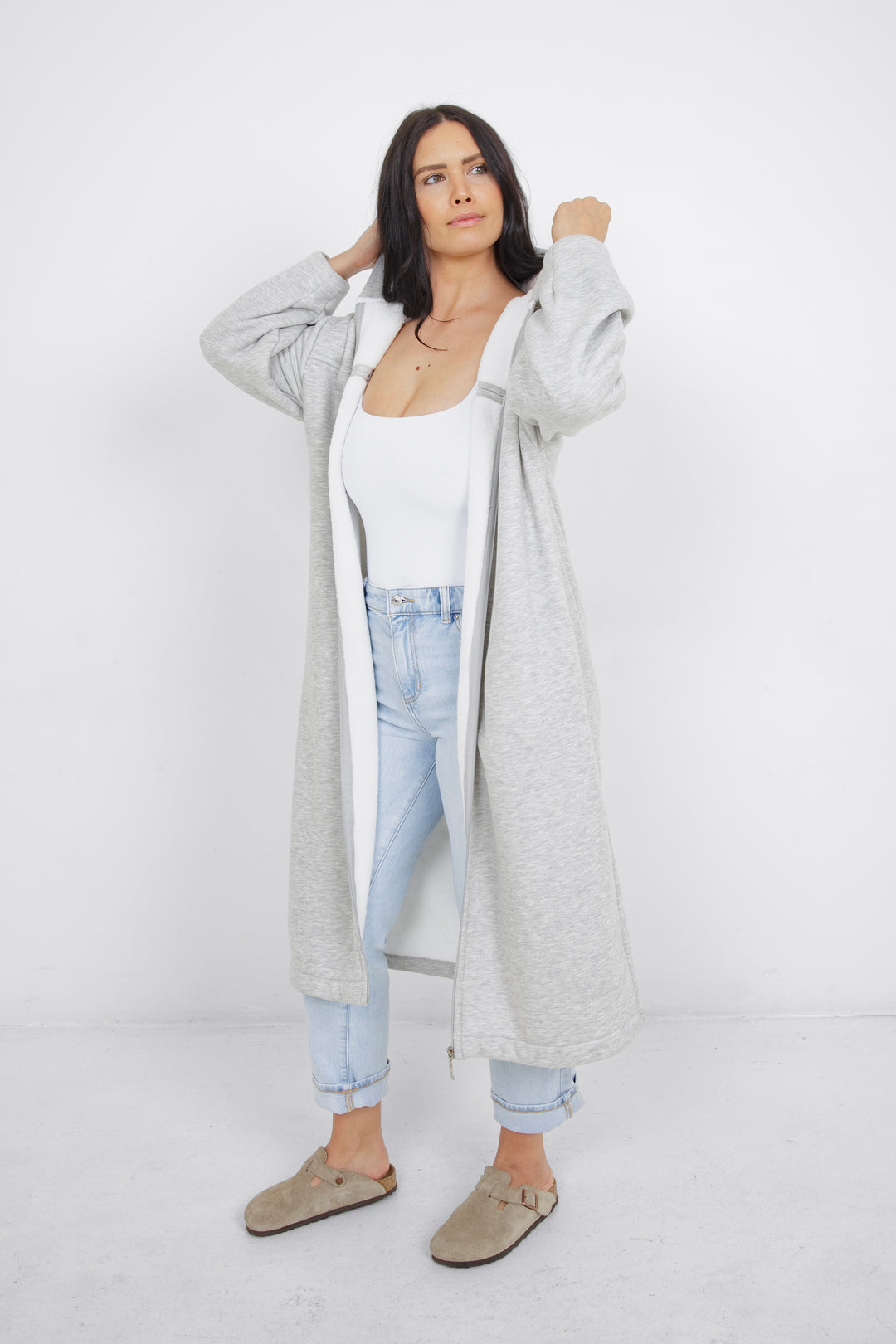 Woman wearing heather grey a long zip up hoodie, unzipped with side slits and pockets in front of a plain grey background