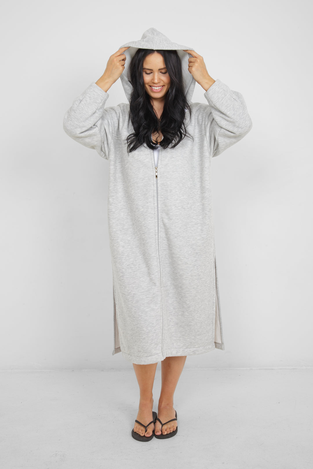 Woman wearing heather grey a long zip up hoodie, zipped up with hood up, with side slits and pockets in front of a plain grey background