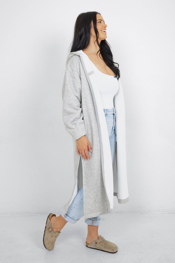 Woman wearing heather grey a long zip up hoodie, unzipped, with side slits and pockets in front of a plain grey background.