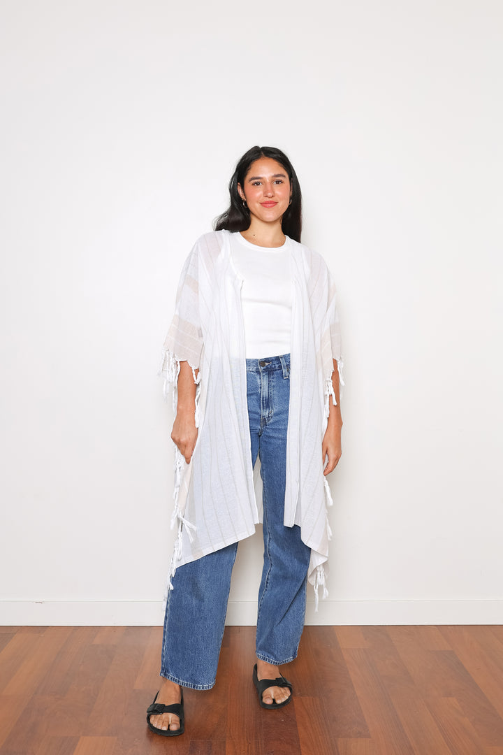 A woman standing wearing a white open cover-up with subtle stripes and tassels, paired with blue jeans and black sandals.