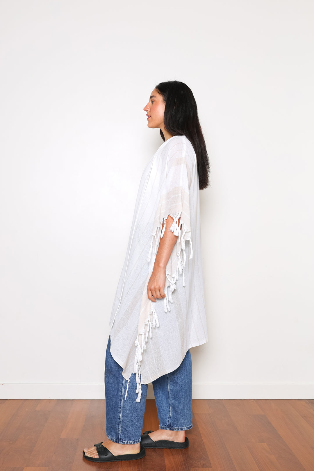 A woman standing wearing a white open cover-up with subtle stripes and tassels, paired with blue jeans and black sandals.