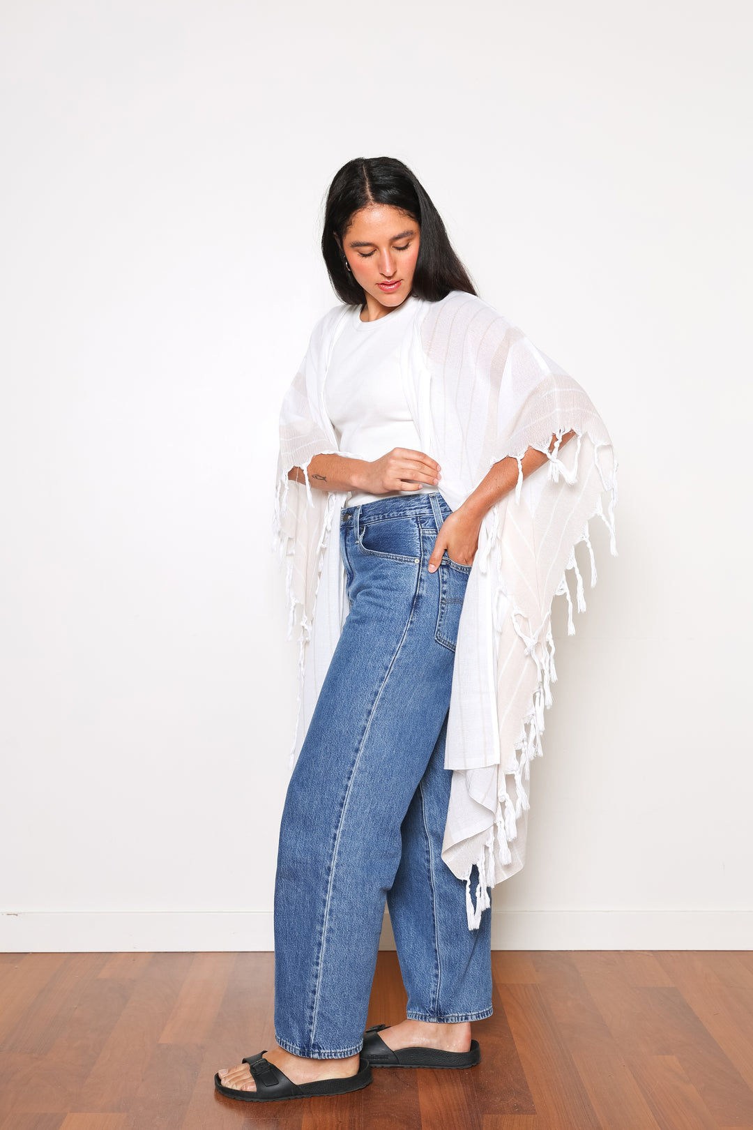 A woman standing wearing a white open cover-up with subtle stripes and tassels, paired with blue jeans and black sandals.