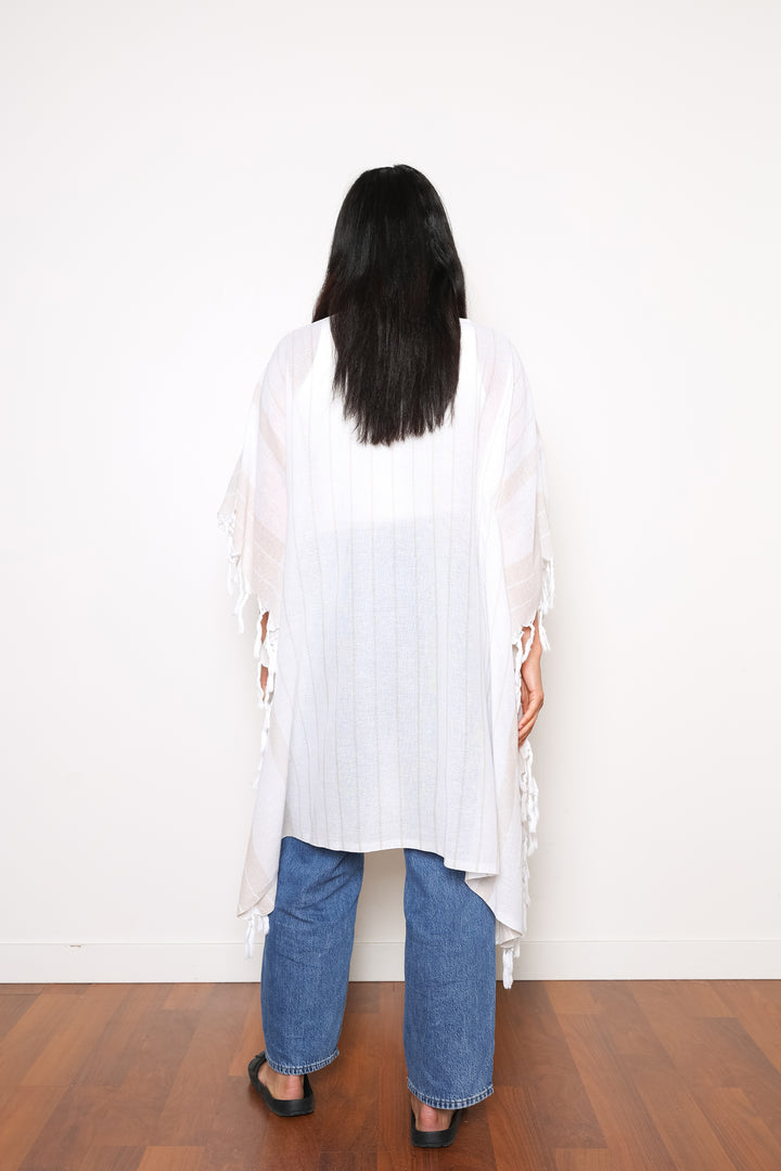 A woman standing backwards wearing a white open cover-up with subtle stripes and tassels, paired with blue jeans and black sandals.