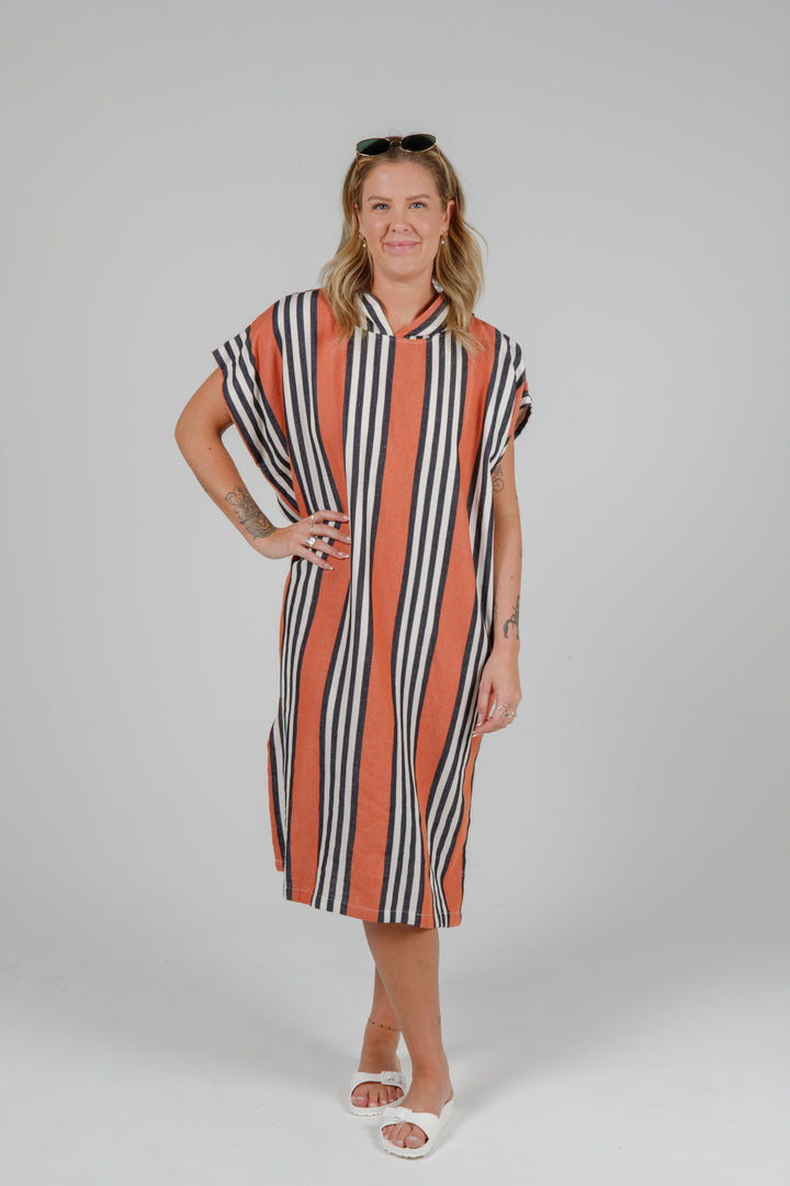 Woman wearing a long warm orange poncho with black and white stripes, side slits and a hood standing in front of a plain light grey background