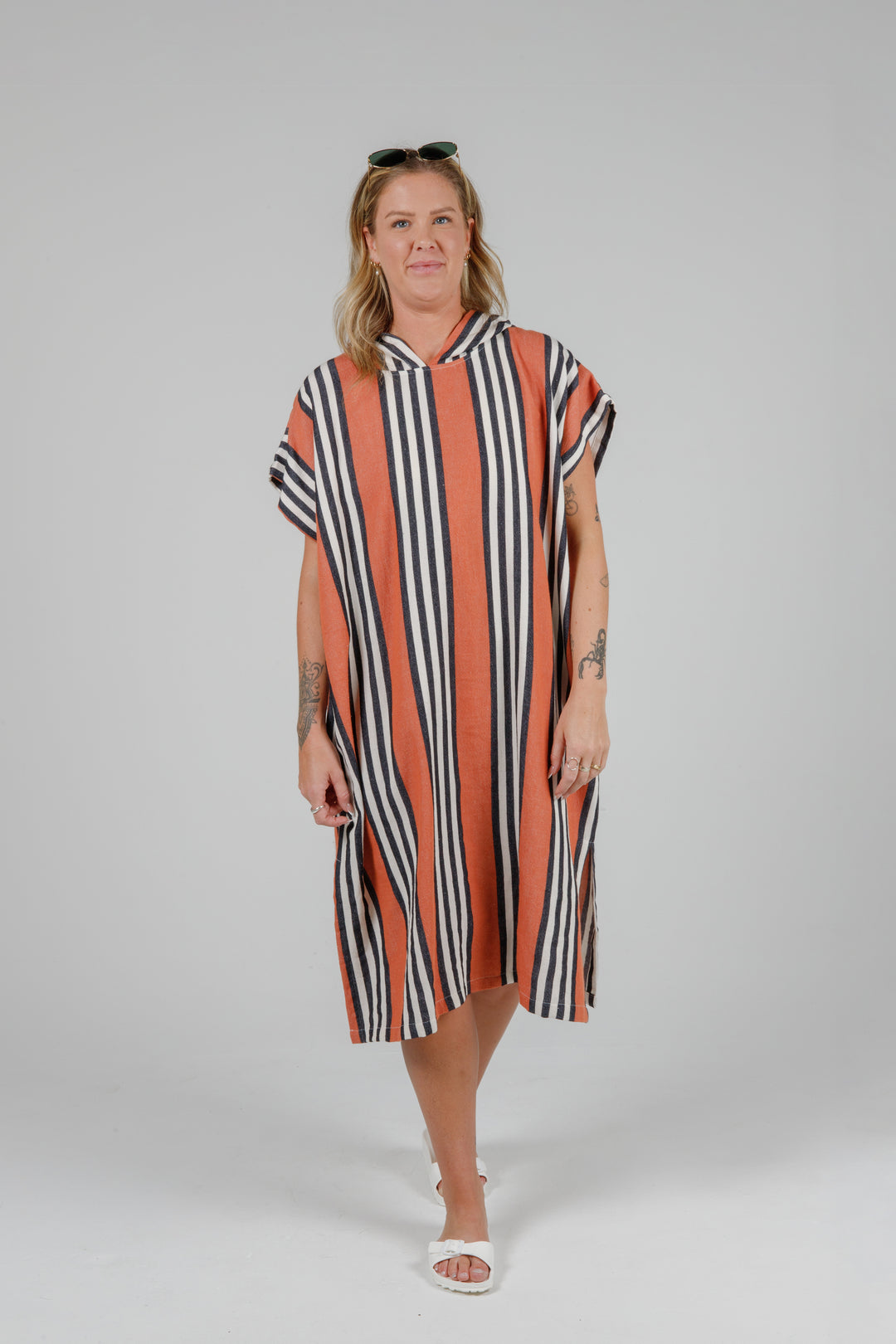 Woman wearing a long warm orange poncho with black and white stripes, side slits and a hood standing in front of a plain light grey background