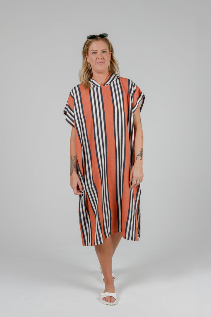 Woman wearing a long warm orange poncho with black and white stripes, side slits and a hood standing in front of a plain light grey background