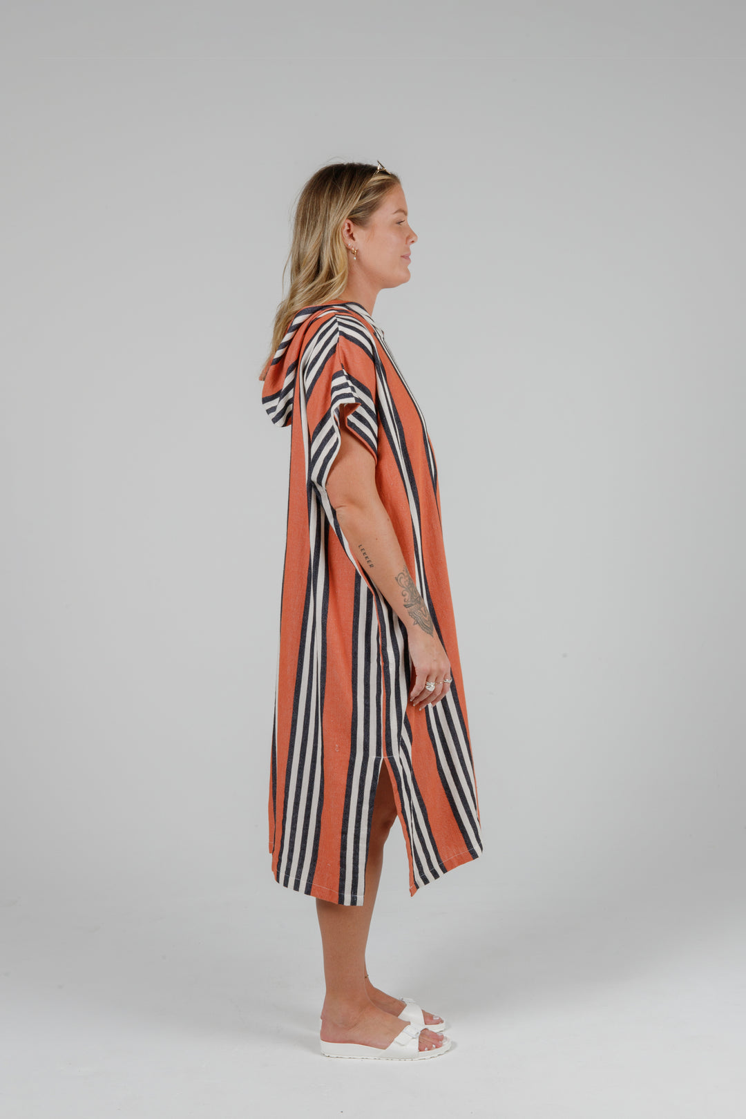 Woman wearing a long warm orange poncho with black and white stripes, side slits and a hood standing in front of a plain light grey background
