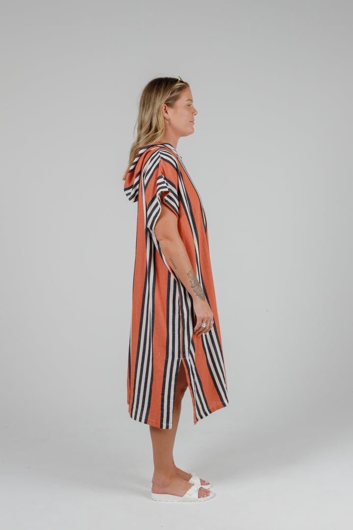 Woman wearing a long warm orange poncho with black and white stripes, side slits and a hood standing in front of a plain light grey background