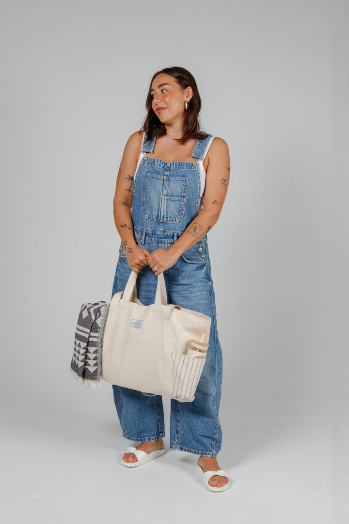 Person wearing denim overalls holding a Large off white Turkish cotton canvas tote bag with exterior pockets, handles containing a geometric towel and a water bottle on a grey background
