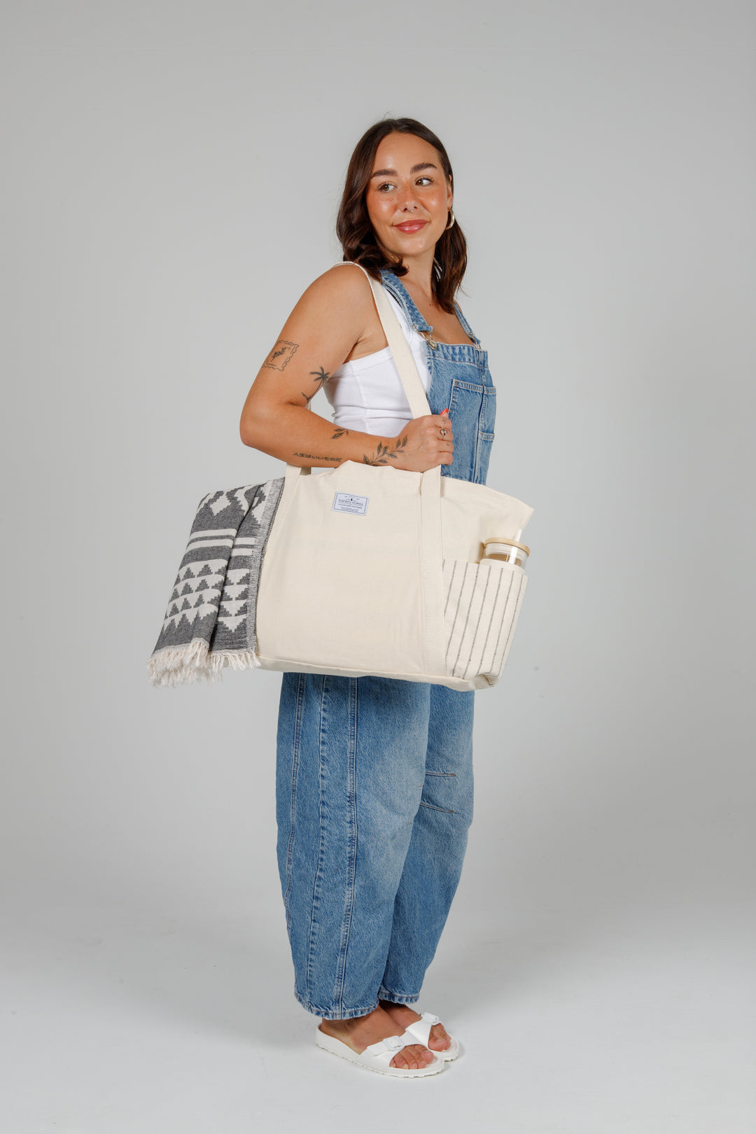 Person wearing denim overalls holding a Large off white Turkish cotton canvas tote bag with exterior pockets, handles containing a geometric towel and a water bottle on a grey background