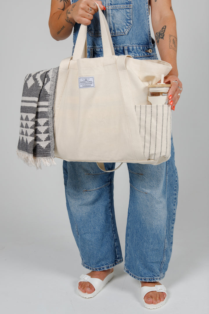 Person wearing denim overalls holding a Large off white Turkish cotton canvas tote bag with exterior pockets and handles containing a geometric towel and a water bottle on a grey background