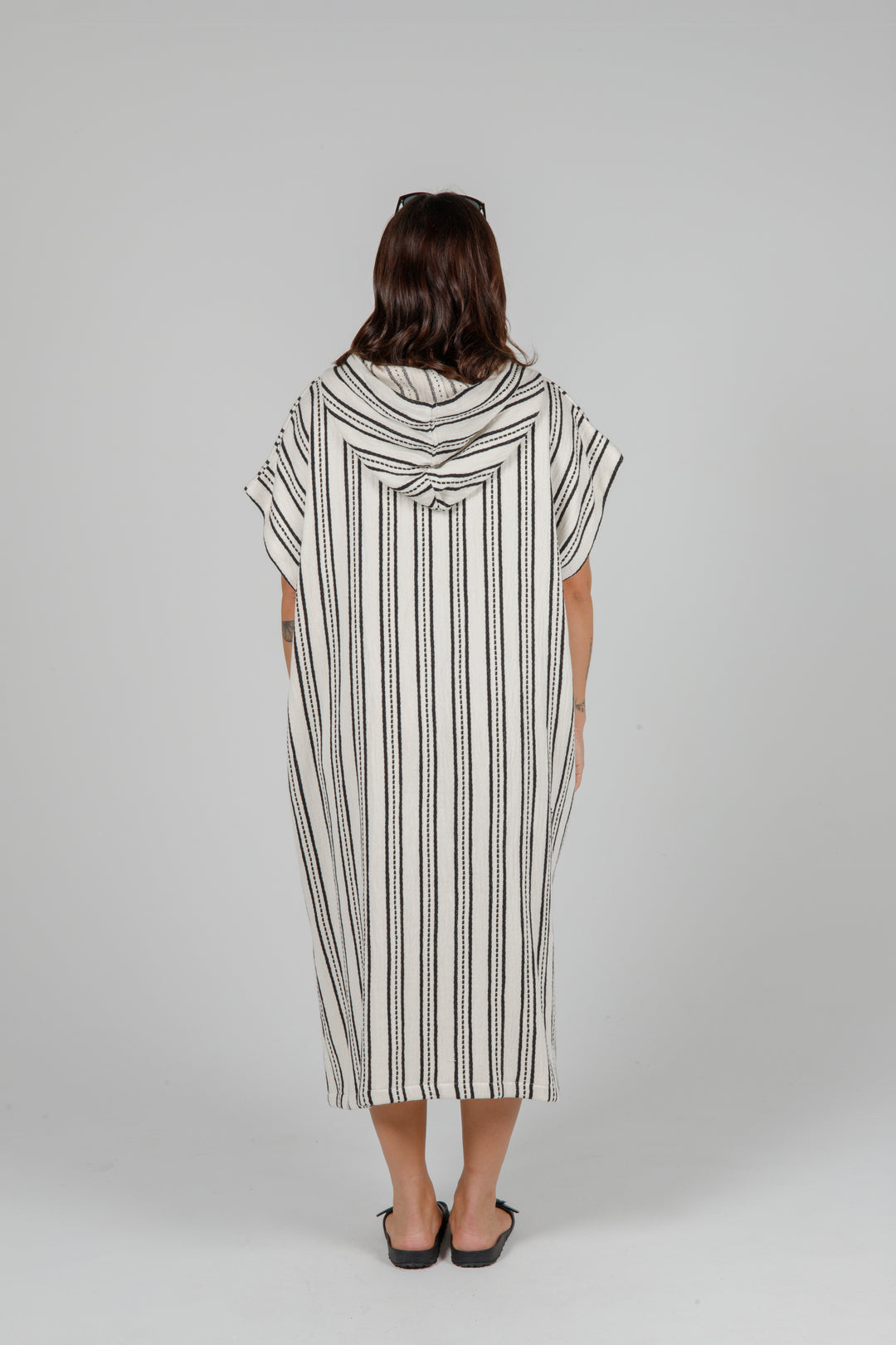 Woman standing backwards wearing a long off white hooded poncho with black hand stitched stripes side slits against a plain background.