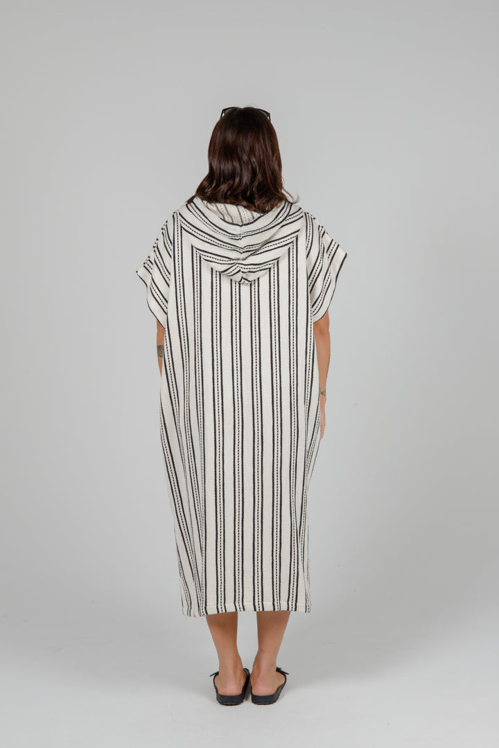 Woman standing backwards wearing a long off white hooded poncho with black hand stitched stripes side slits against a plain background.