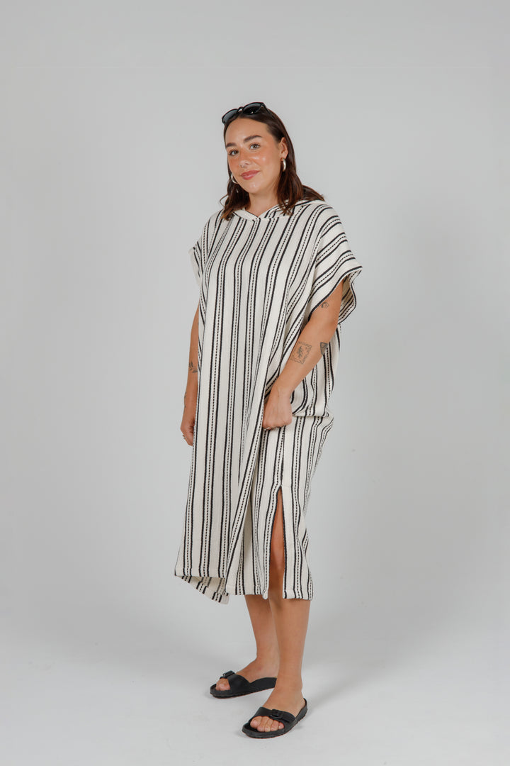 Woman wearing a long off white hooded poncho with black hand stitched stripes side slits against a plain background.