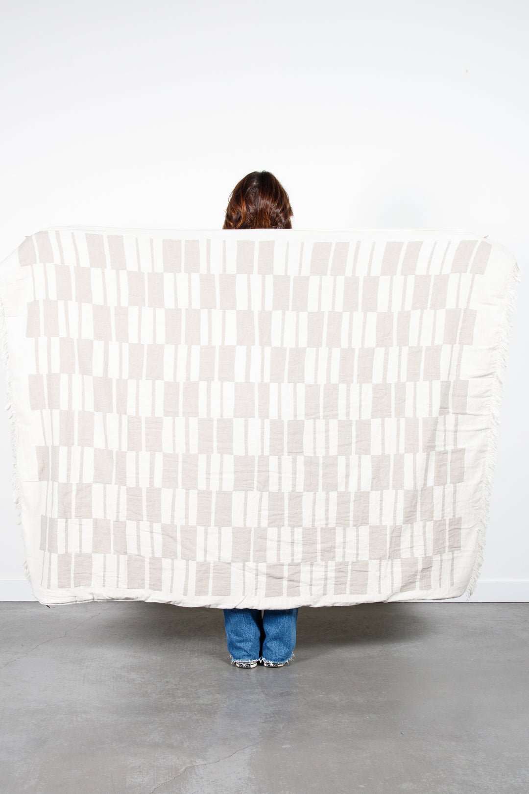 woman standing backwards holding up an off white and beige fleece lined throw with modern checker board pattern and eyelash fringe behind her #colour_beige