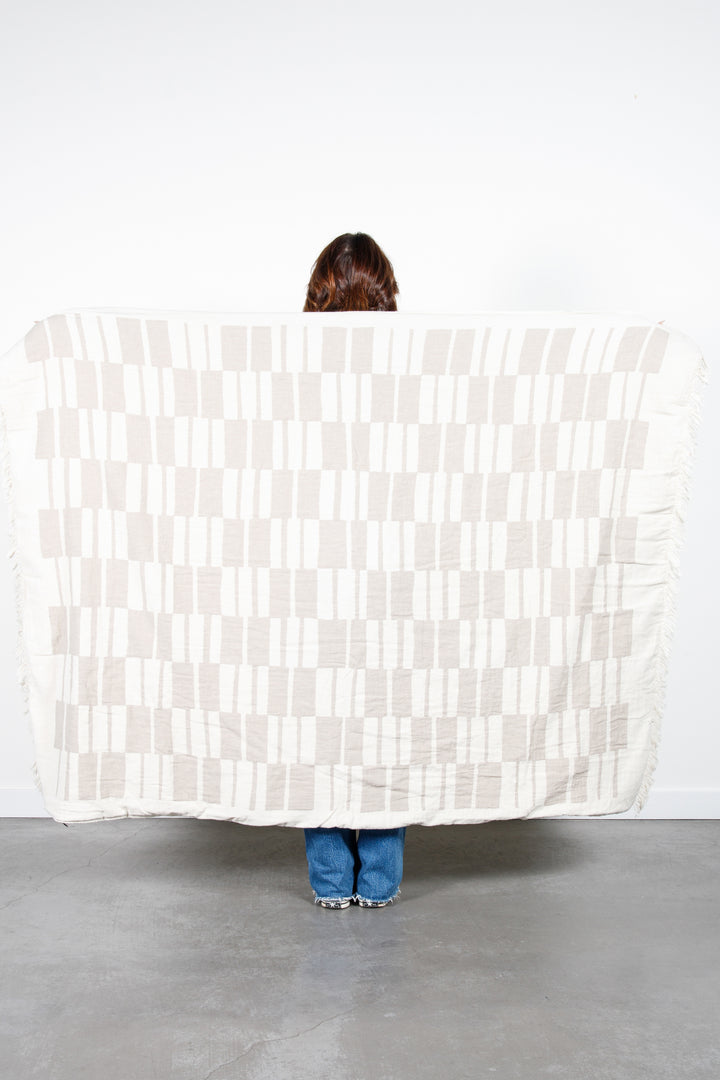 woman standing backwards holding up an off white and beige fleece lined throw with modern checker board pattern and eyelash fringe behind her #colour_beige