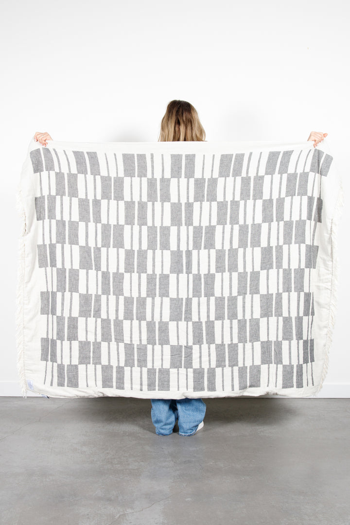 woman standing backwards holding up an off white and dark grey fleece lined throw with modern checker board pattern and eyelash fringe behind her #colour_granite