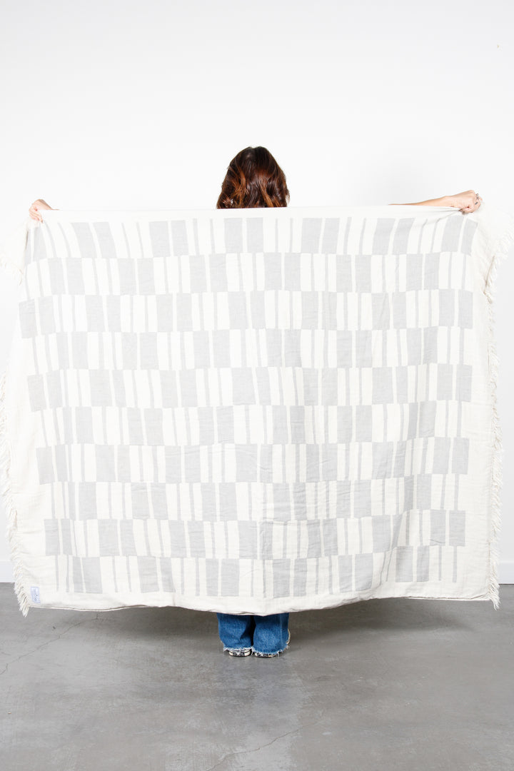 woman standing backwards holding up an off white and light grey fleece lined throw with modern checker board pattern and eyelash fringe behind her #colour_pewter