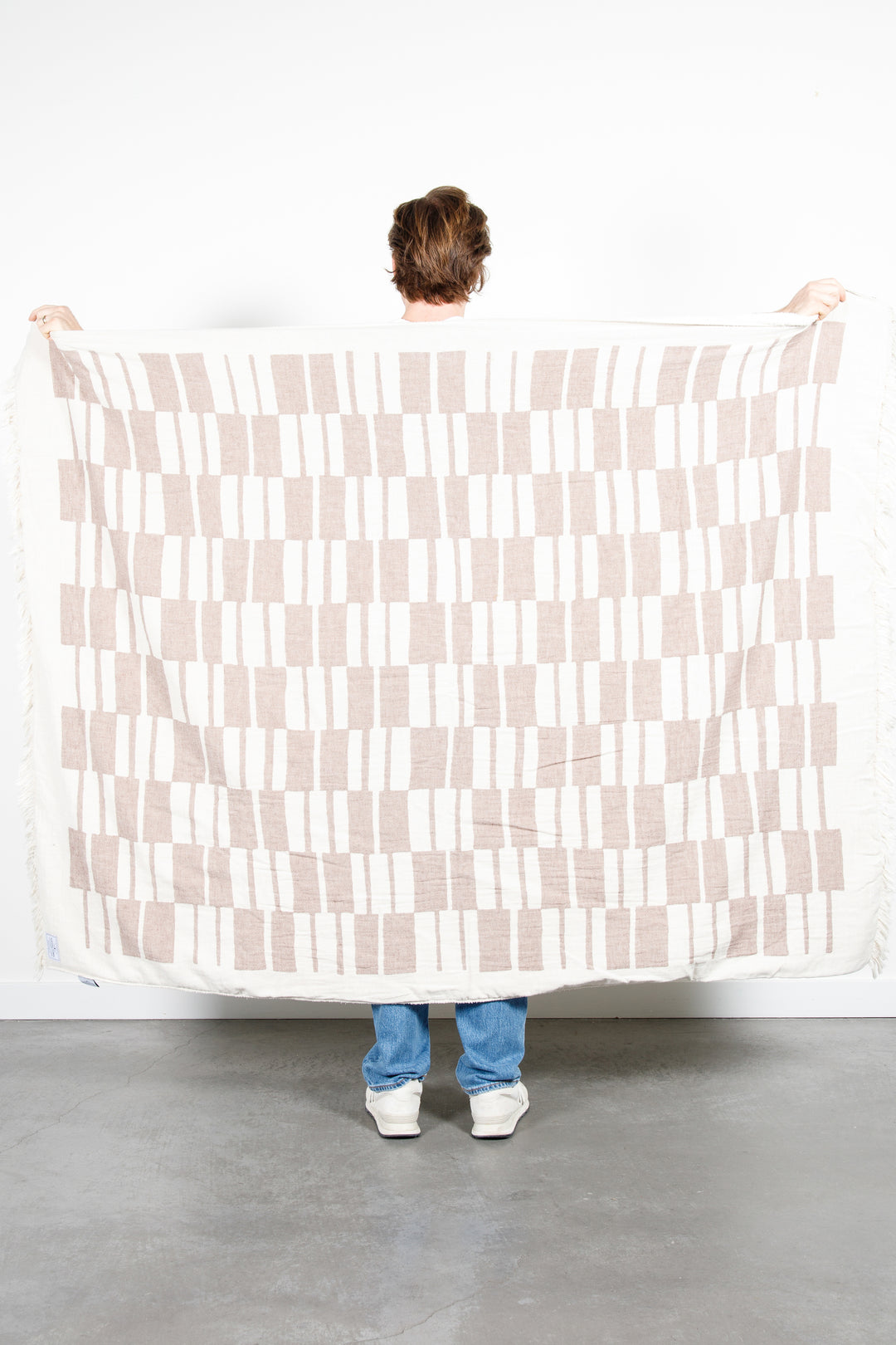 woman standing backwards holding up an off white and light brown fleece lined throw with modern checker board pattern and eyelash fringe behind her #colour_walnut