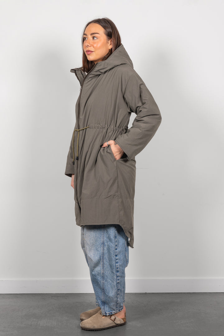 A woman modeling a mid-length olive green hooded parka jacket with a drawstring waist, paired with light blue jeans and brown clogs, standing against a plain light gray wall.