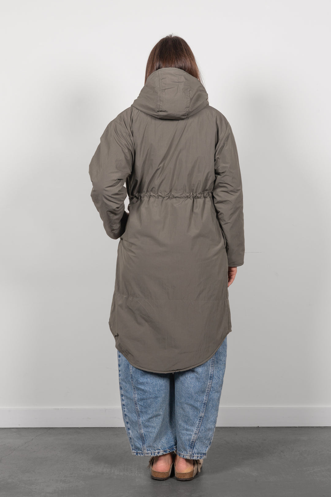 A person viewed from the back wearing a long hooded puffer coat in a muted taupe-gray color, paired with loose blue jeans and brown shoes, standing against a plain light gray wall.