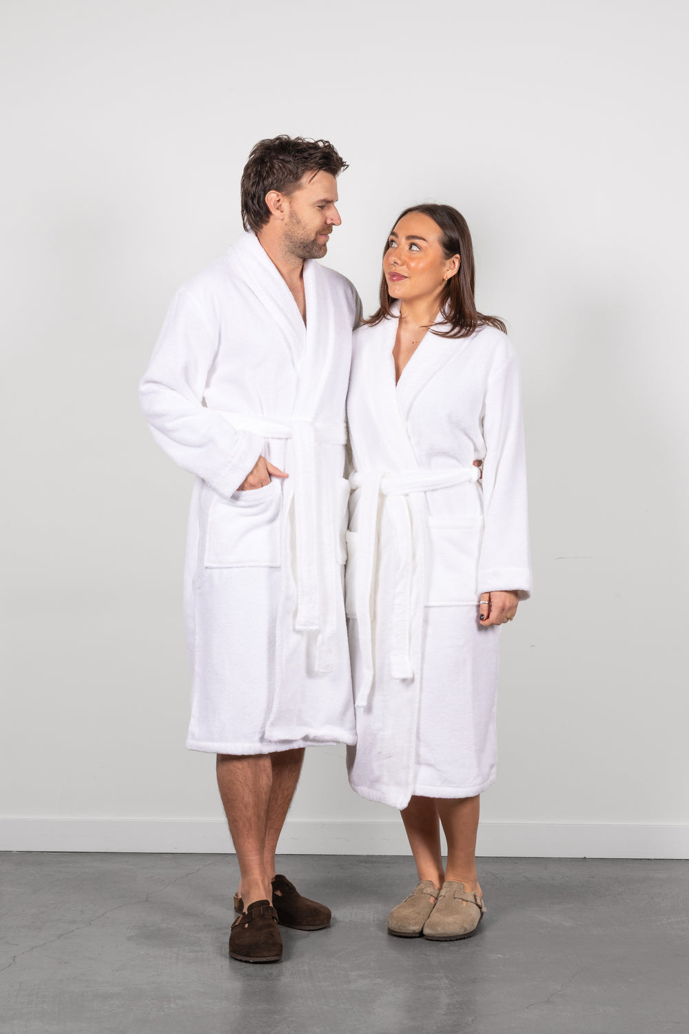 A man and a woman standing side by side wearing matching white bathrobes and slippers, photographed against a plain light gray wall and gray floor. #colour_off-white